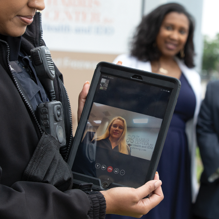 A Houston Police Officer is holding a tablet device toward the viewer. They are conducting a video call with a member of the Harris Team.