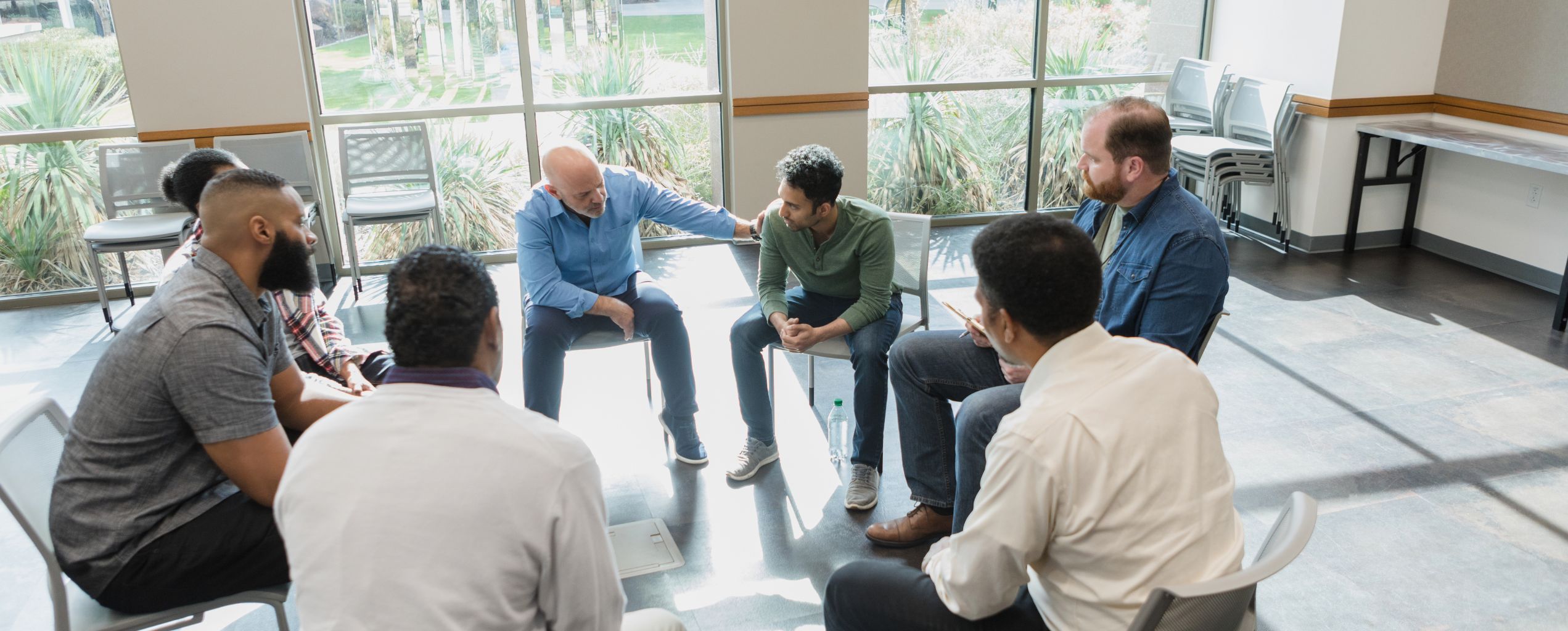 A support group of diverse men sitting in a circle, with one man offering comfort to another in a sunlit room with large windows.
