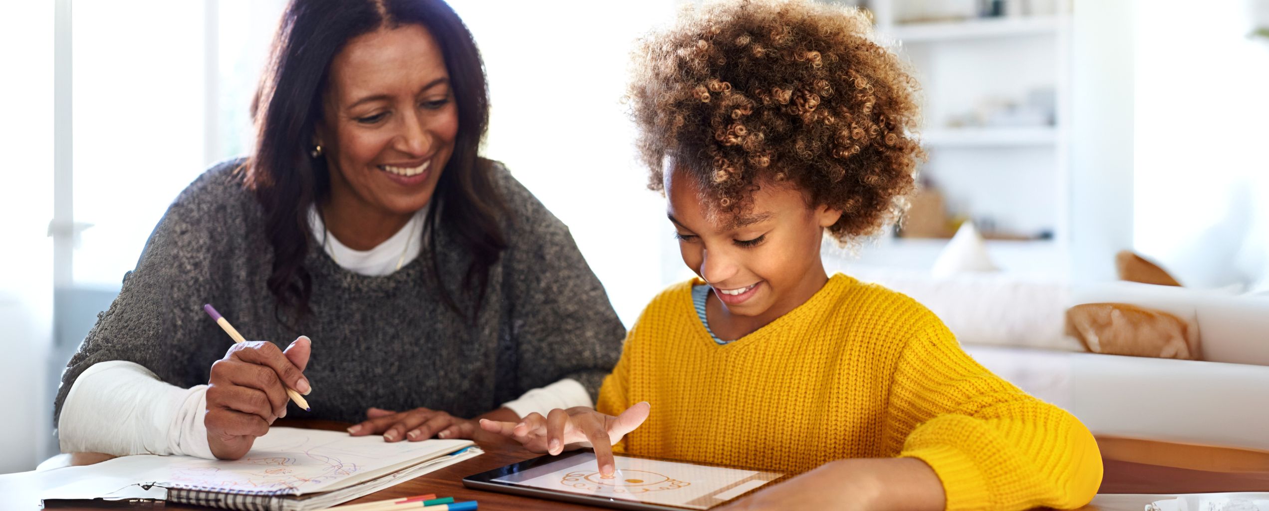 "Smiling woman and child working together on drawings, using a tablet and sketchpad in a bright home setting.