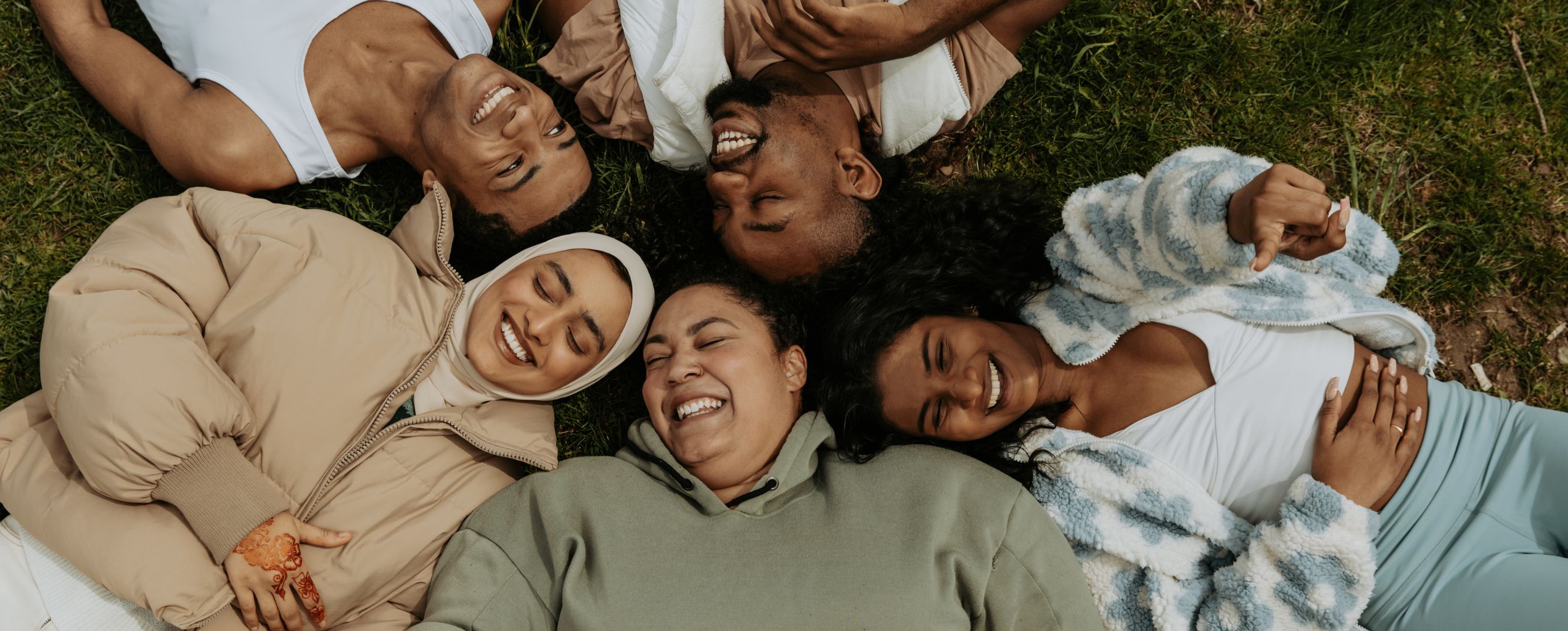 Group of diverse young adults lying on the grass in a circle, smiling and laughing together.