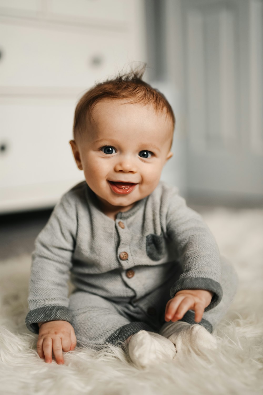 Smiling baby in a gray onesie sitting on a soft white rug indoors.