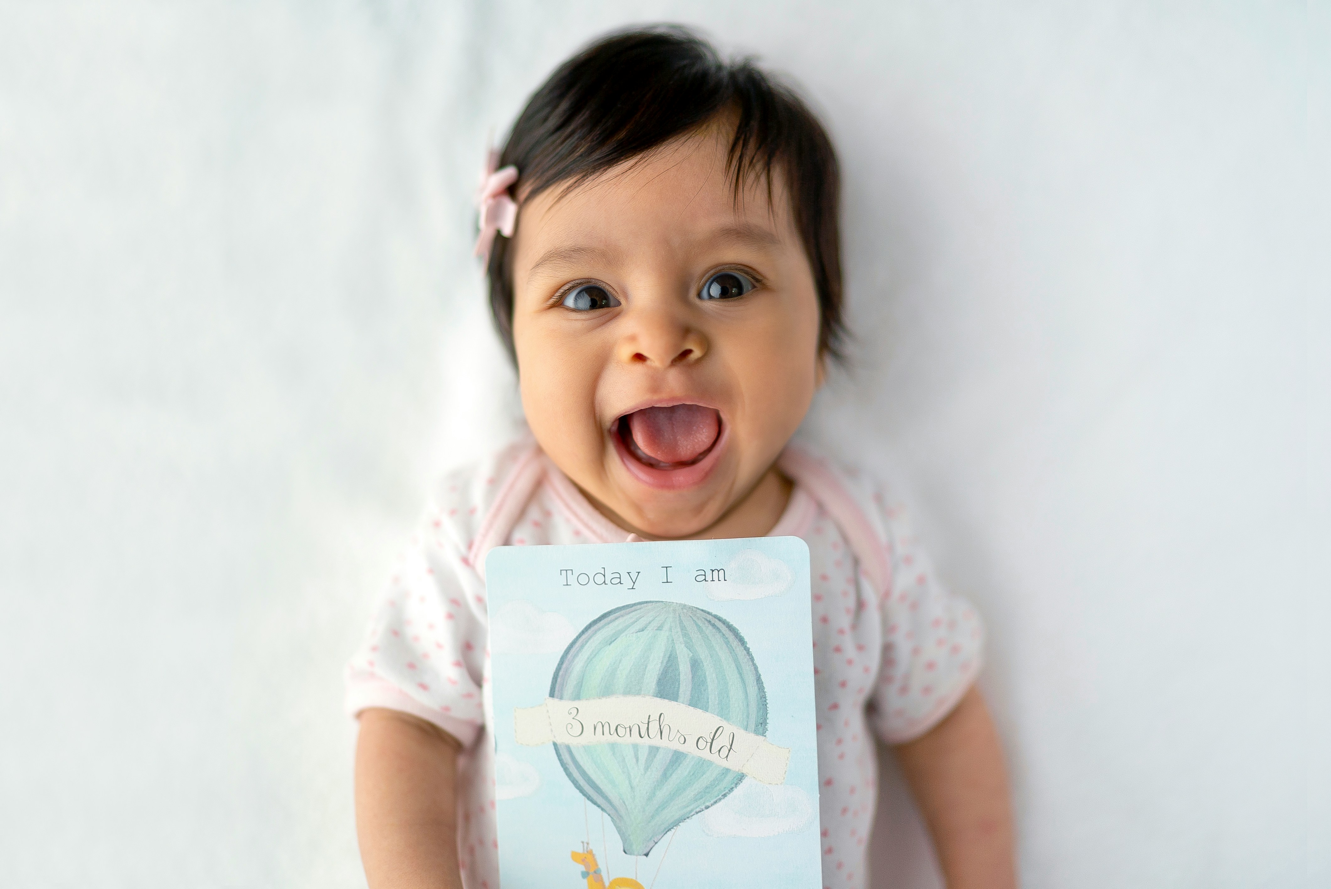 Happy baby in a pink onesie lying on a white blanket, holding a milestone card that reads Today I am 3 months old.