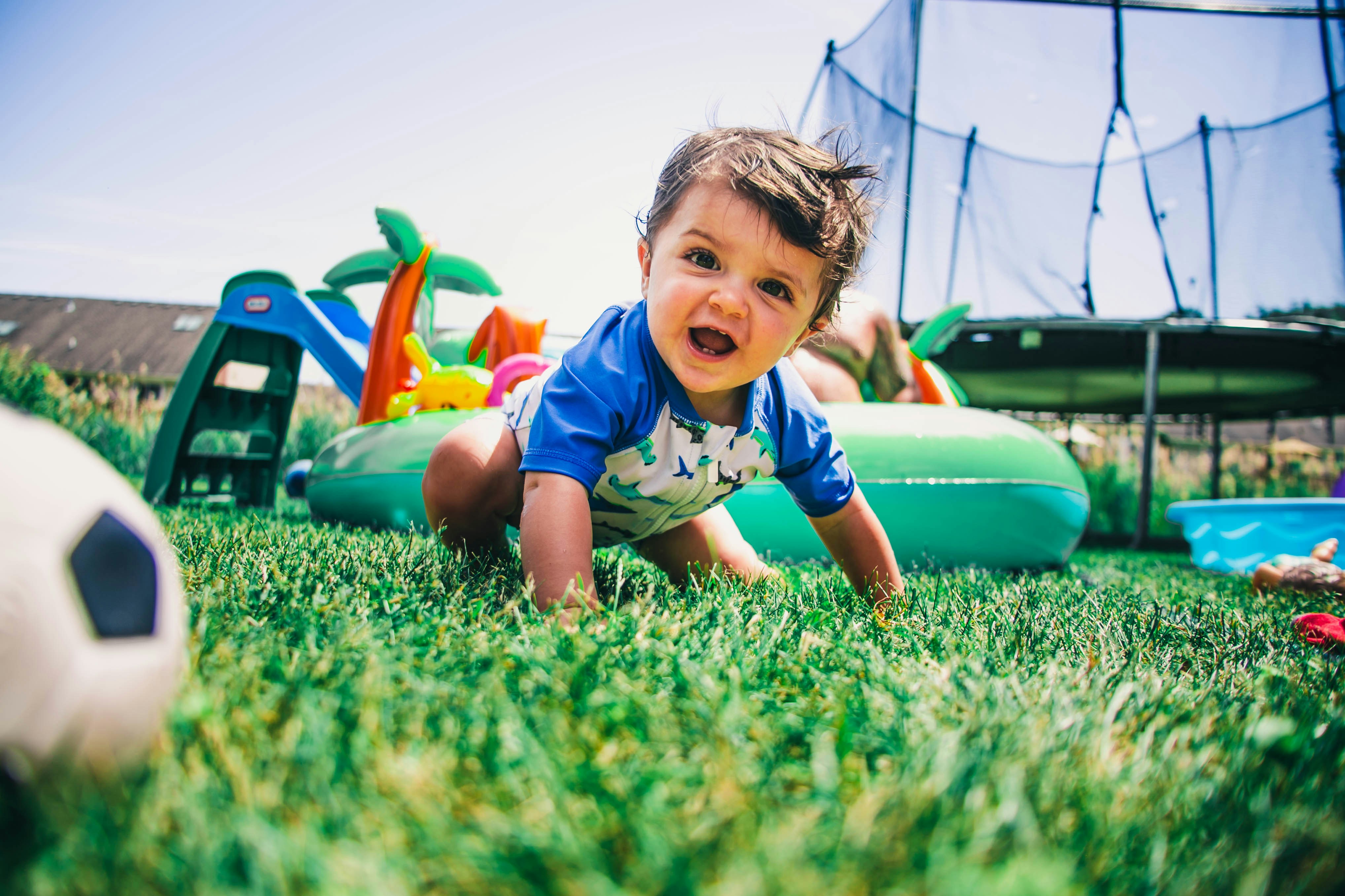 Smiling baby crawling on green grass in a backyard, wearing a blue swimsuit, with a slide, inflatable pool, and trampoline in the background.