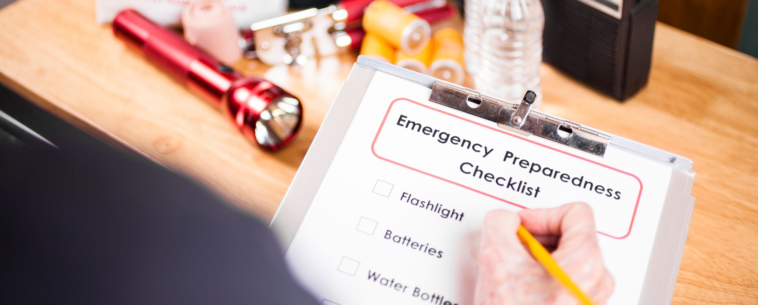 Close-up of a person writing on an emergency preparedness checklist with items like flashlight, batteries, and water bottles, alongside supplies on a table.