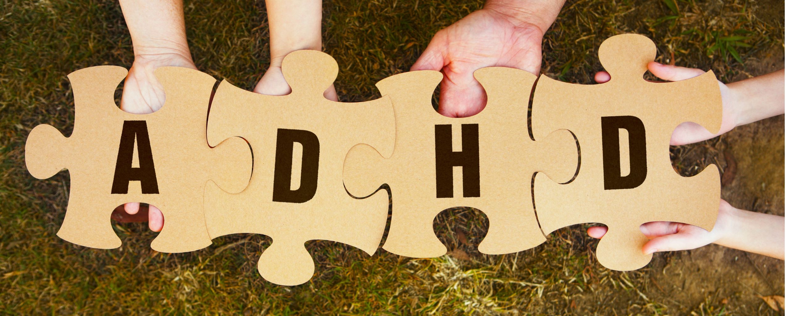 Four hands hold four large puzzle pieces that fit together to spell “ADHD,” symbolizing awareness and connection. The puzzle pieces are brown cardboard against a grassy background.