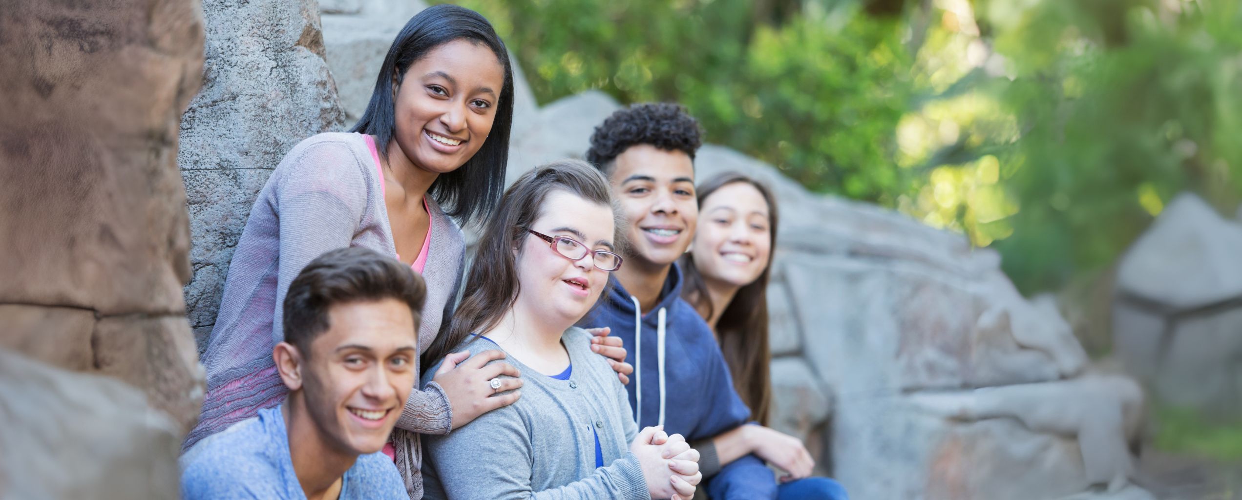 A diverse group of smiling teenagers sitting together outdoors on stone steps, including a young woman with Down syndrome in the center, symbolizing inclusion and friendship.