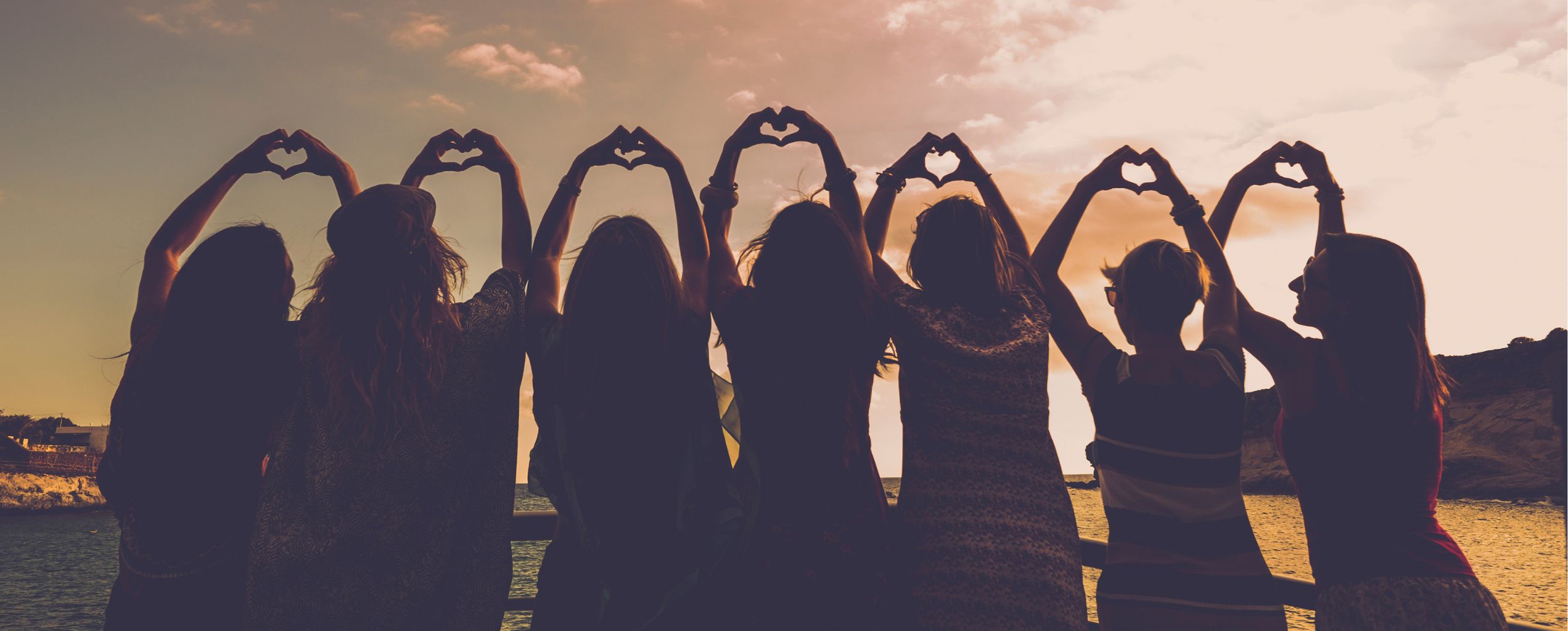 A group of people standing in a row at sunset, raising their arms to form heart shapes with their hands, symbolizing unity and friendship.