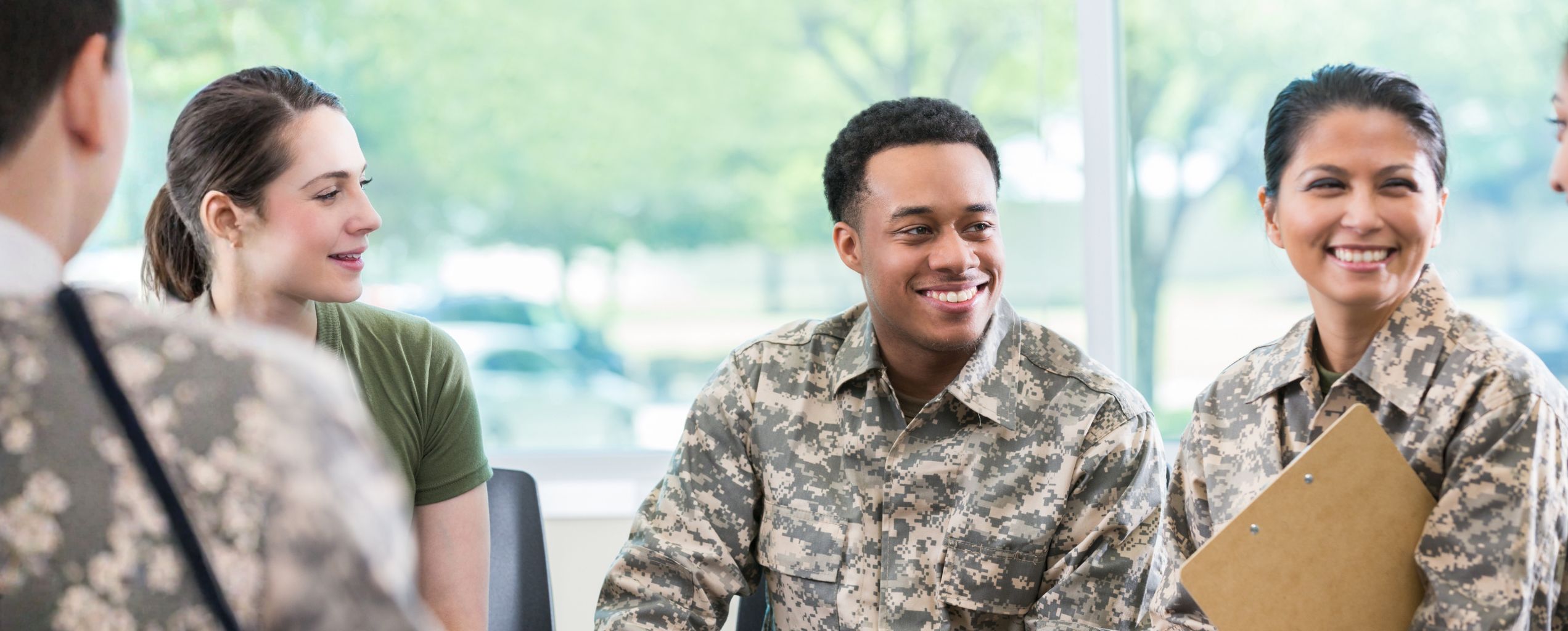 A group of smiling military service members in uniform sitting together indoors, engaged in friendly conversation.