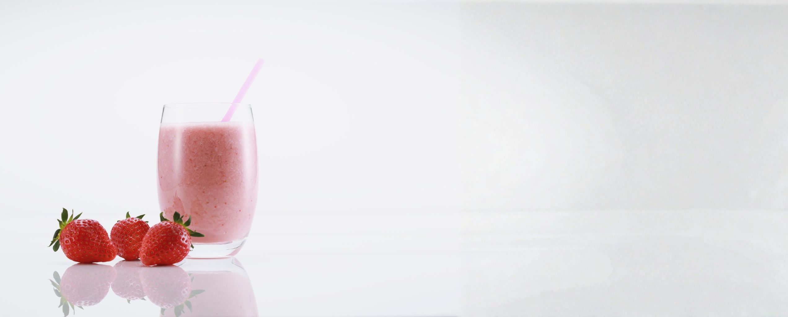 “Glass of pink strawberry smoothie with a straw, placed on a white surface beside fresh strawberries, with a clean minimalist background.
