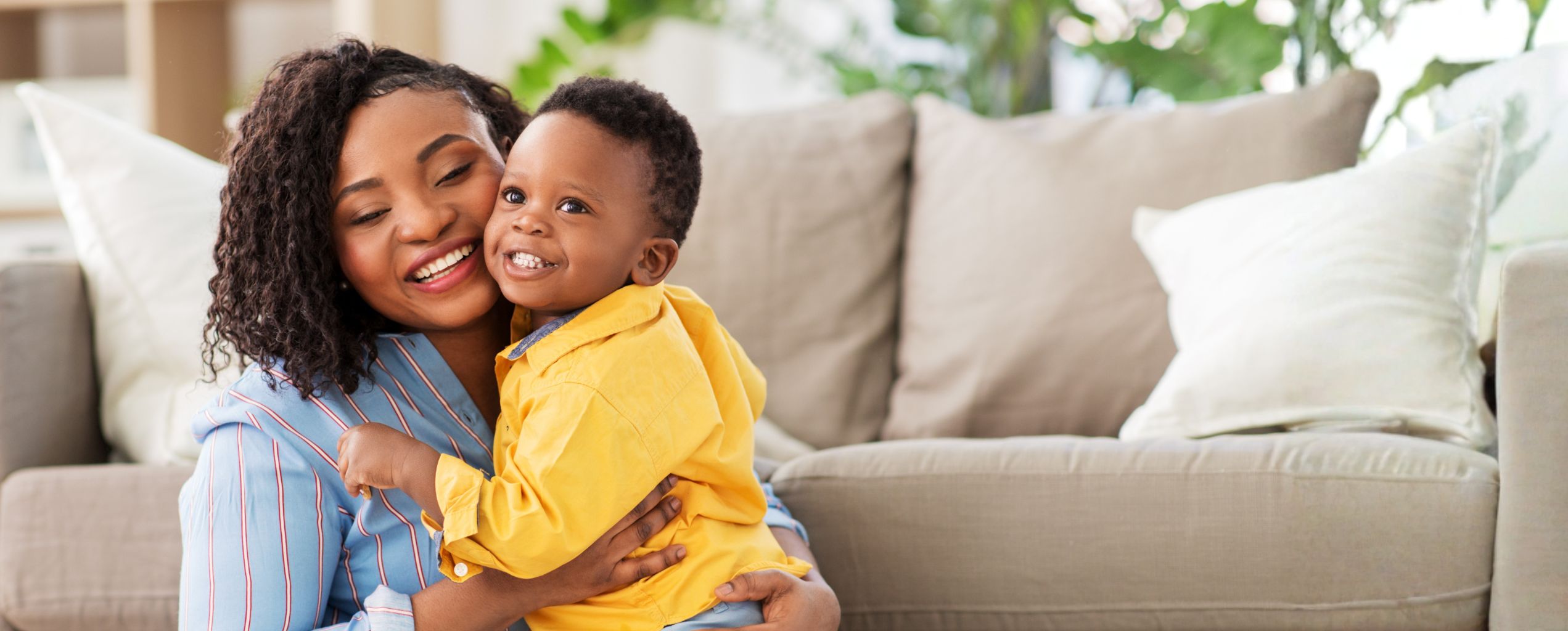 Smiling mother holding her young child in a bright living room, both looking happy and close while sitting on the floor in front of a couch.