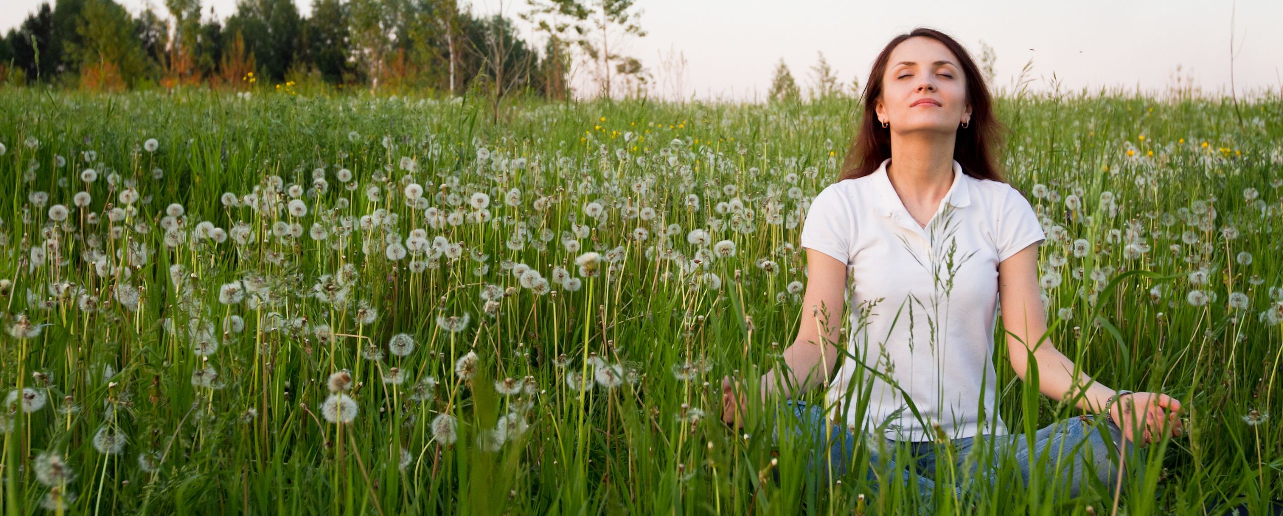 Woman sitting cross-legged in a green meadow filled with dandelions, eyes closed and hands resting on her knees, appearing calm and relaxed while enjoying nature.