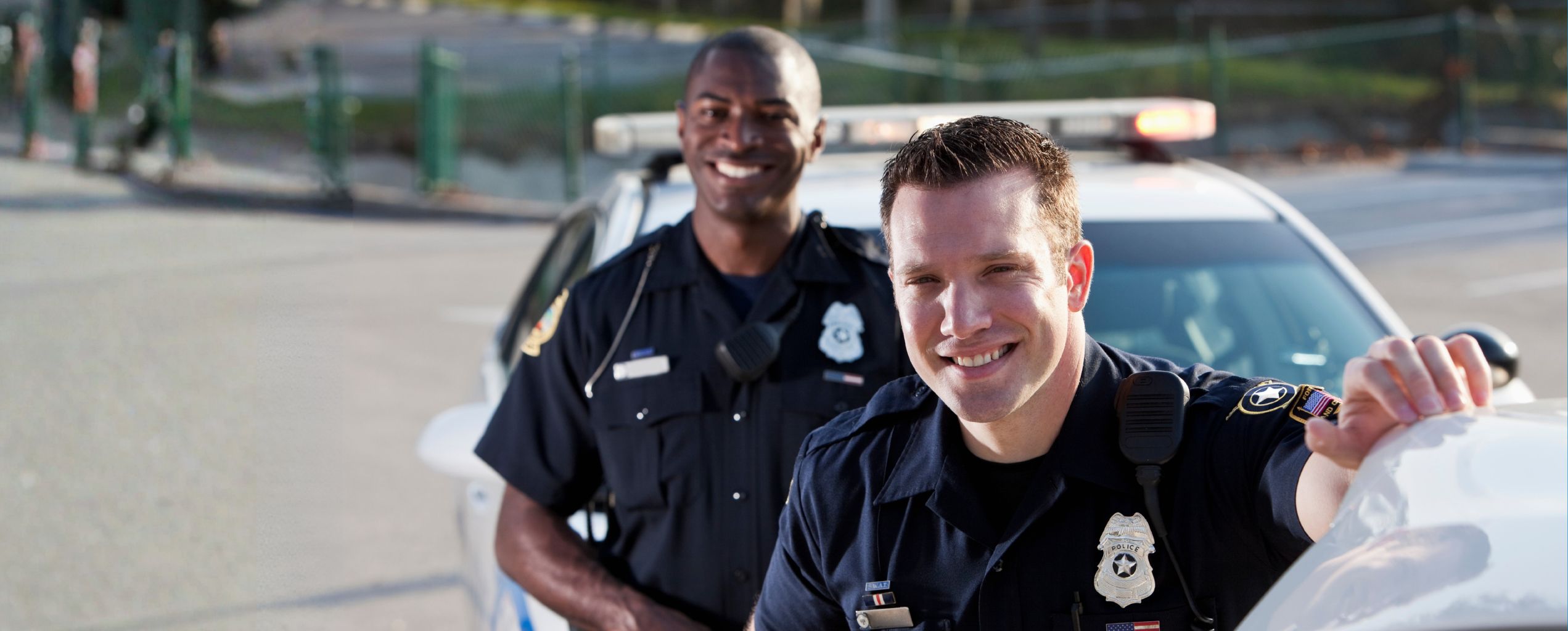 Two uniformed police officers smiling in front of a patrol car outdoors, with one officer leaning on the vehicle and the other standing behind.