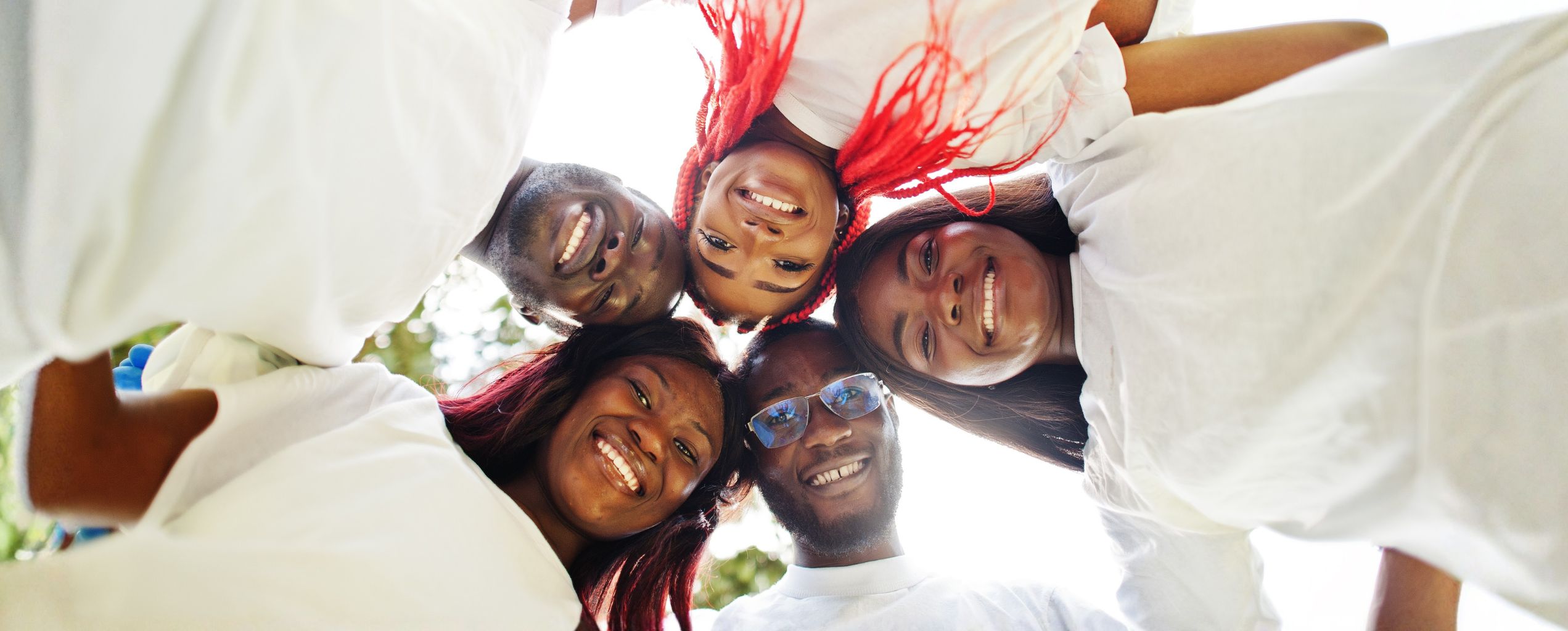 Group of six people smiling and looking down at the camera while standing in a circle outdoors.