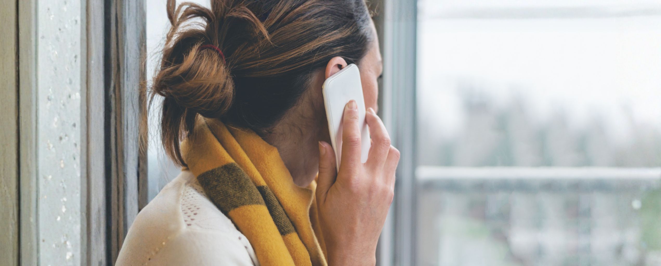 Person standing by a window, holding a phone to their ear while looking outside, appearing thoughtful and engaged in a call.