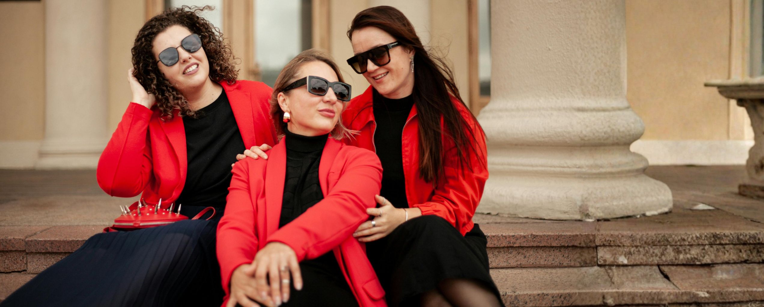 Three women wearing red jackets and black outfits sit together on stone steps, smiling and posing confidently outdoors.