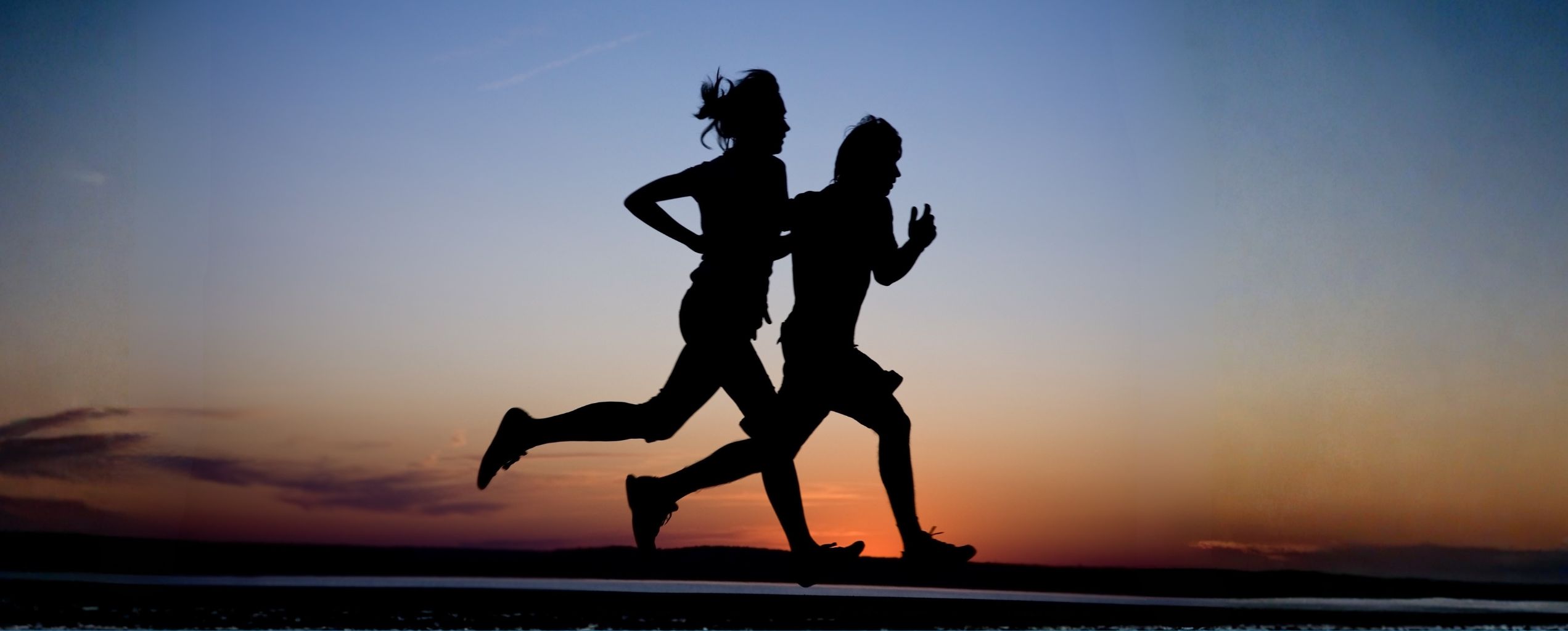 Silhouettes of two people running side by side at sunset, outlined against a colorful evening sky.