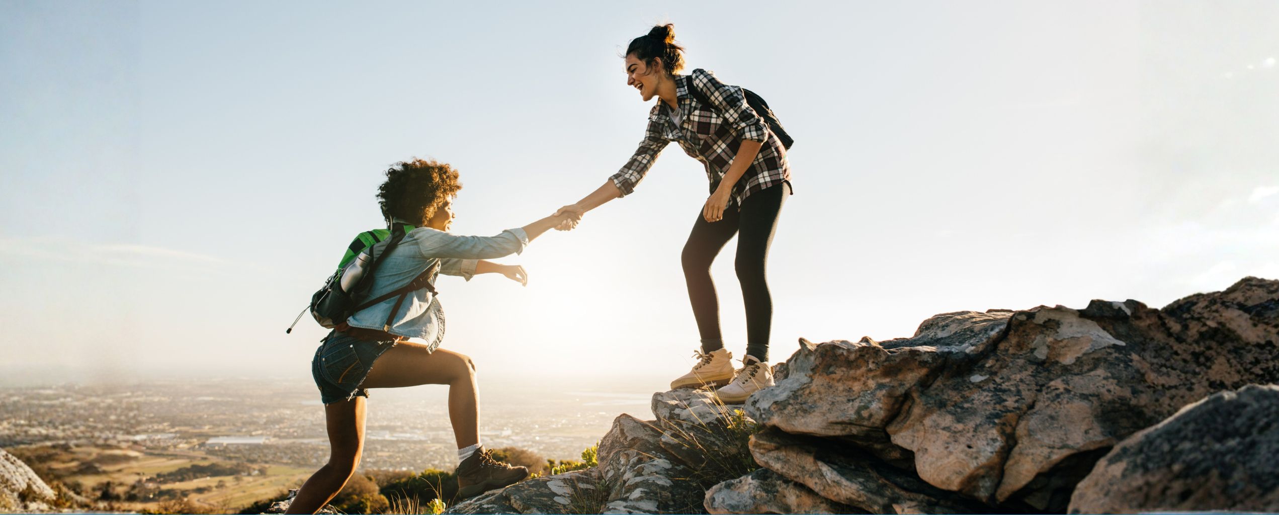 A woman helping her hiking companion climb up a rocky mountain slope in bright sunshine.