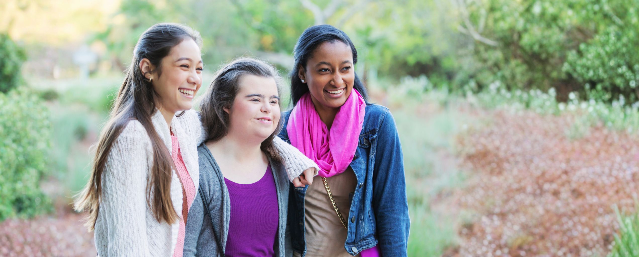 Three women smiling together outdoors surrounded by nature.