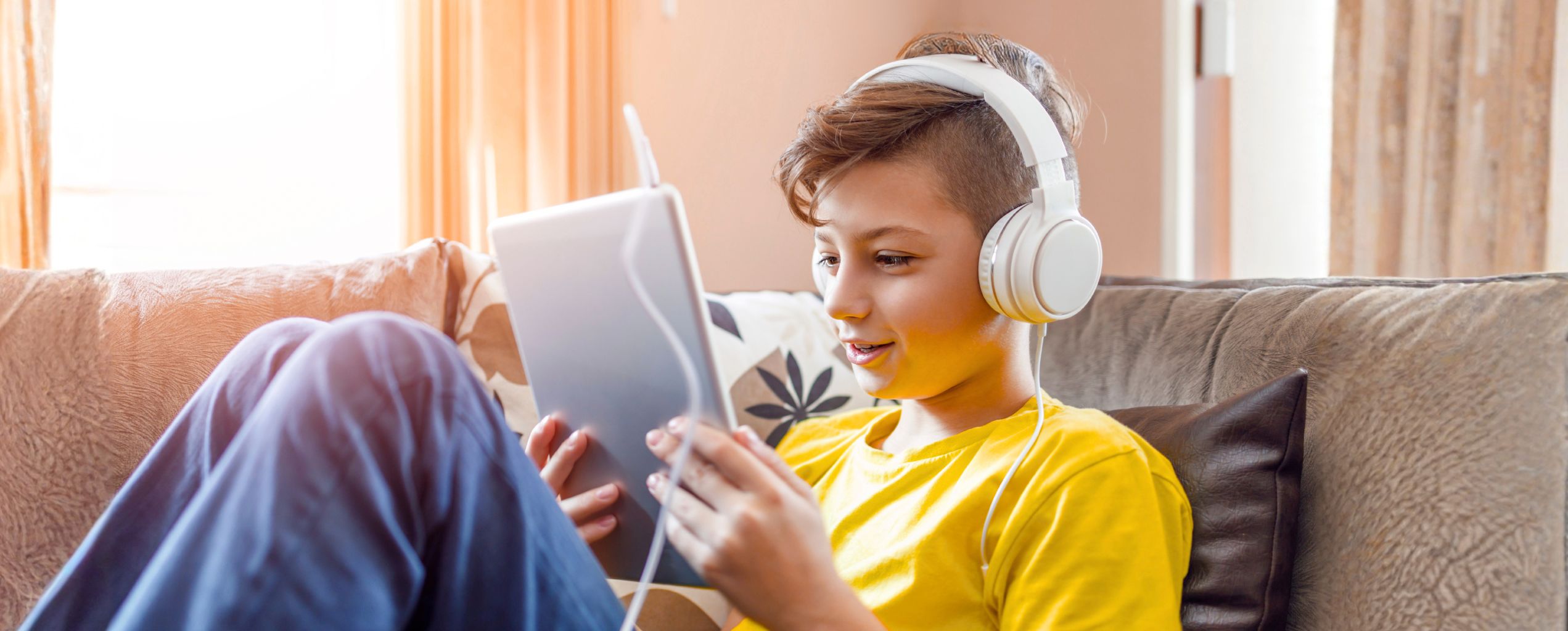 A child sitting on a couch in a sunlit room, listening to music and using a tablet.