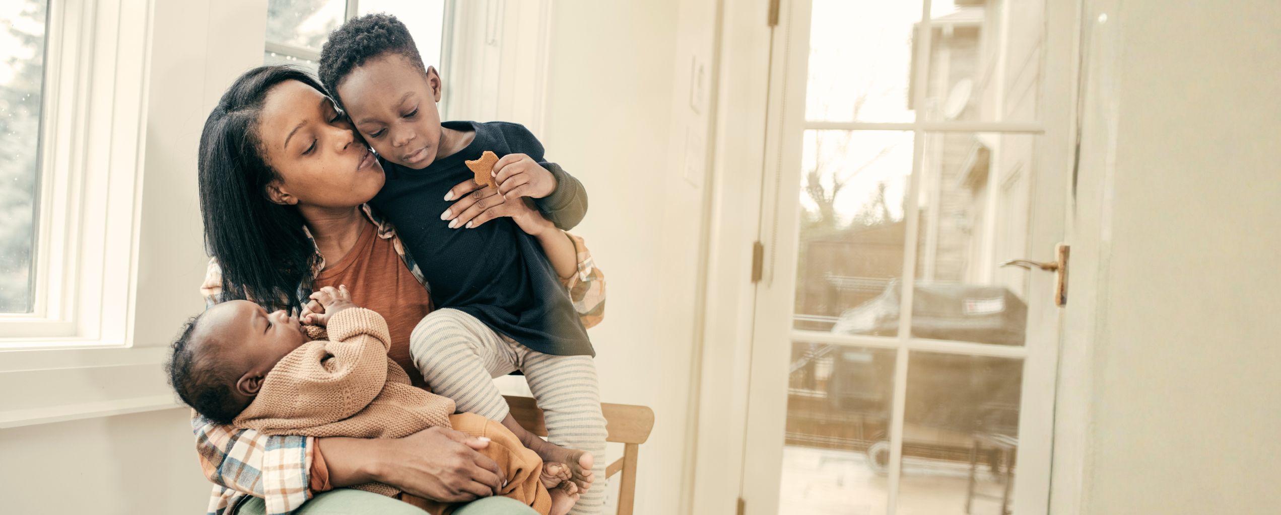 Mother holding her baby and toddler in a bright, sunlit room.