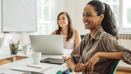 Two business professionals sitting at a shared desk in a bright office, smiling at an unseen speaker